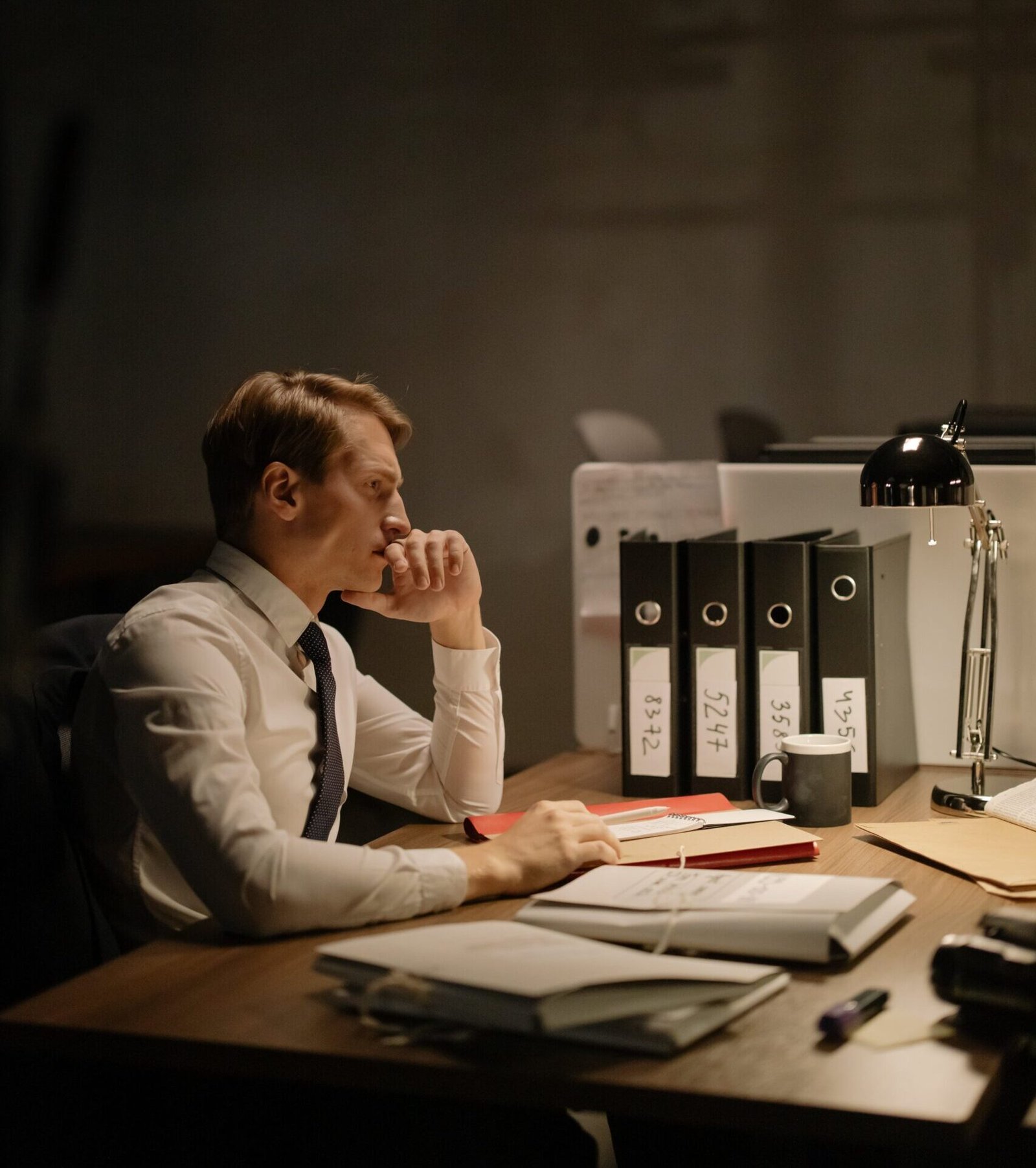 Thoughtful Man in White Shirt and Black Tie Sitting at a Desk with File Folders